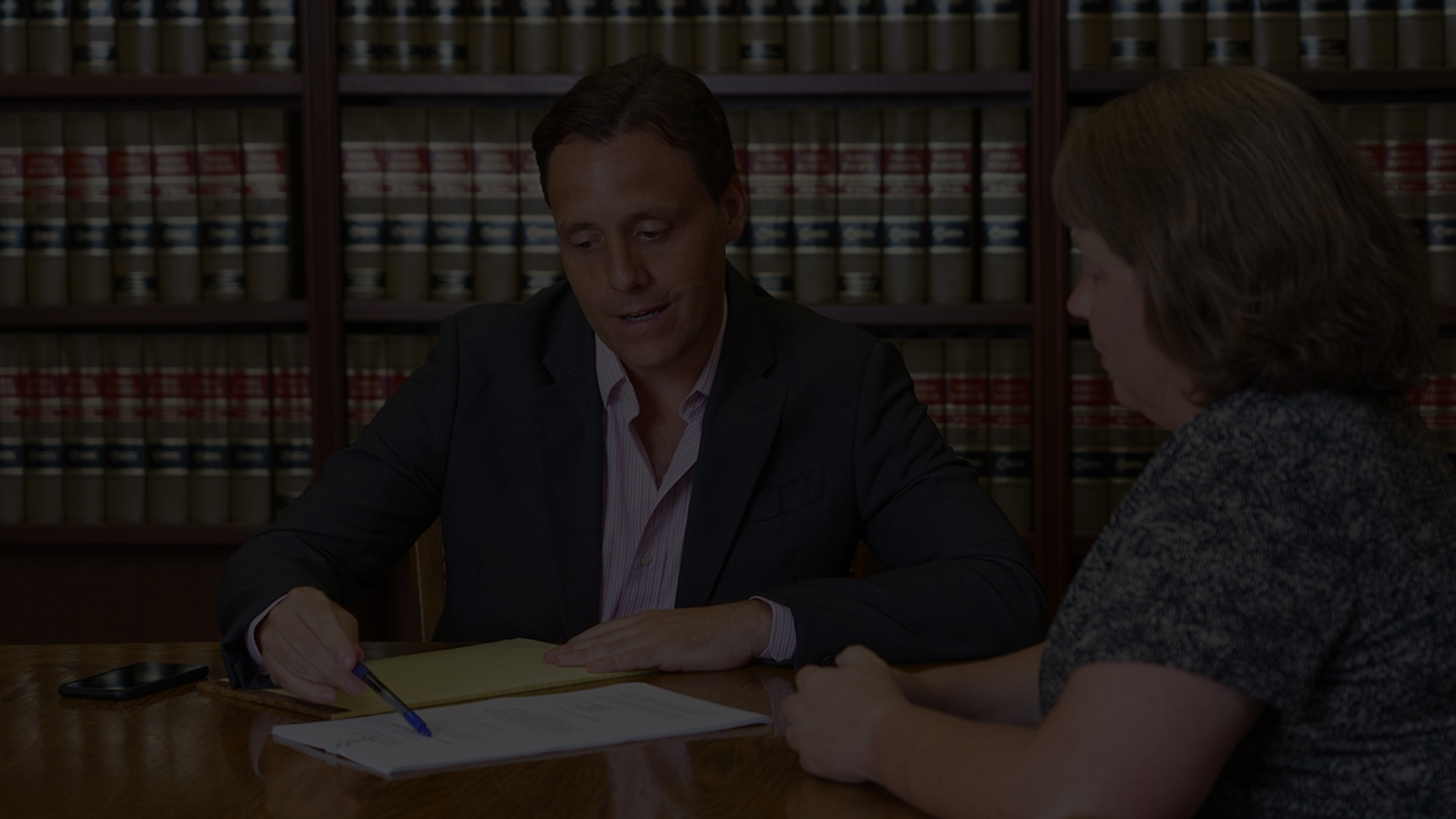 A man in a suit explains documents to a woman at a table, with shelves of legal books in the background.
